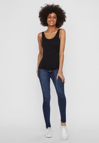 Black sleeveless tank top paired with dark blue slim-fit jeans and white sneakers. Model stands with hands on hips, smiling.