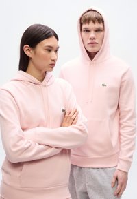 Απαλά ροζ φούτερ με κουκούλα, κατασκευασμένα από μαλακό υλικό, με μικρό πράσινο λογότυπο στο στήθος. Το μοντέλο ποζάρει με τα χέρια σταυρωμένα.