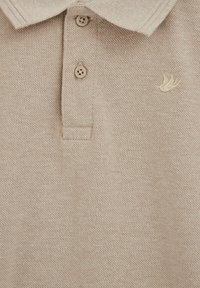 Polo in maglia beige con due bottoni e un piccolo emblema ricamato sul petto a sinistra.