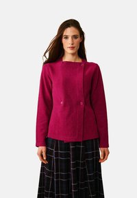 Fuksia villasekoitteinen takki, jossa on pitkät hihat, neliömäinen pääntie ja kaksi etupainiketta. Yhdistetty tumman ruudullisen hameen kanssa, jossa on sinisiä yksityiskohtia.