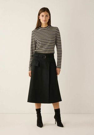 Frau, die steht und ein schwarz-weiß gestreiftes Langarmshirt, einen schwarzen Midirock mit Gürtel und Tasche sowie schwarze hochhackige Stiefel trägt.