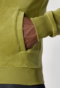 Olive green sweatshirt with a soft texture, ribbed cuffs, and a side pocket. Visible stitching detail at the hem.