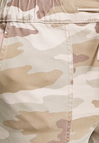 Tissu camouflage beige avec des taches irrégulières en marron clair et kaki. Présente des coutures visibles et une texture de ceinture élastique.