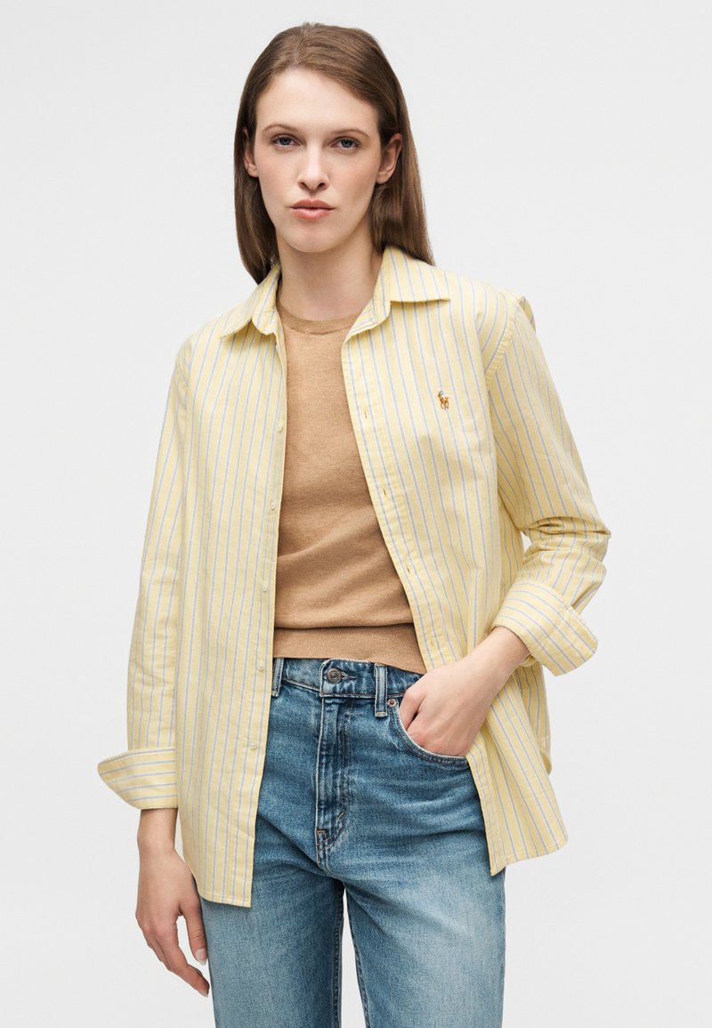 Chemise à rayures jaunes et blanches à boutons, manches retroussées, portée sur un haut marron clair et un jean bleu taille haute.