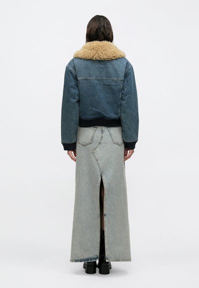 Άτομο που φοράει denim σακάκι με γιακά από ταμπά shearling, μακριά φούστα από ανοιχτό γκρι denim με σκίσιμο πίσω και μαύρα μποτάκια αστραγάλου, στραμμένο προς τα πίσω.