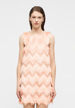 Vrouw draagt een mouwloze perzikkleurige jurk met een textuurrijke zigzagfranjes, staande tegen een effen lichtgrijze achtergrond.