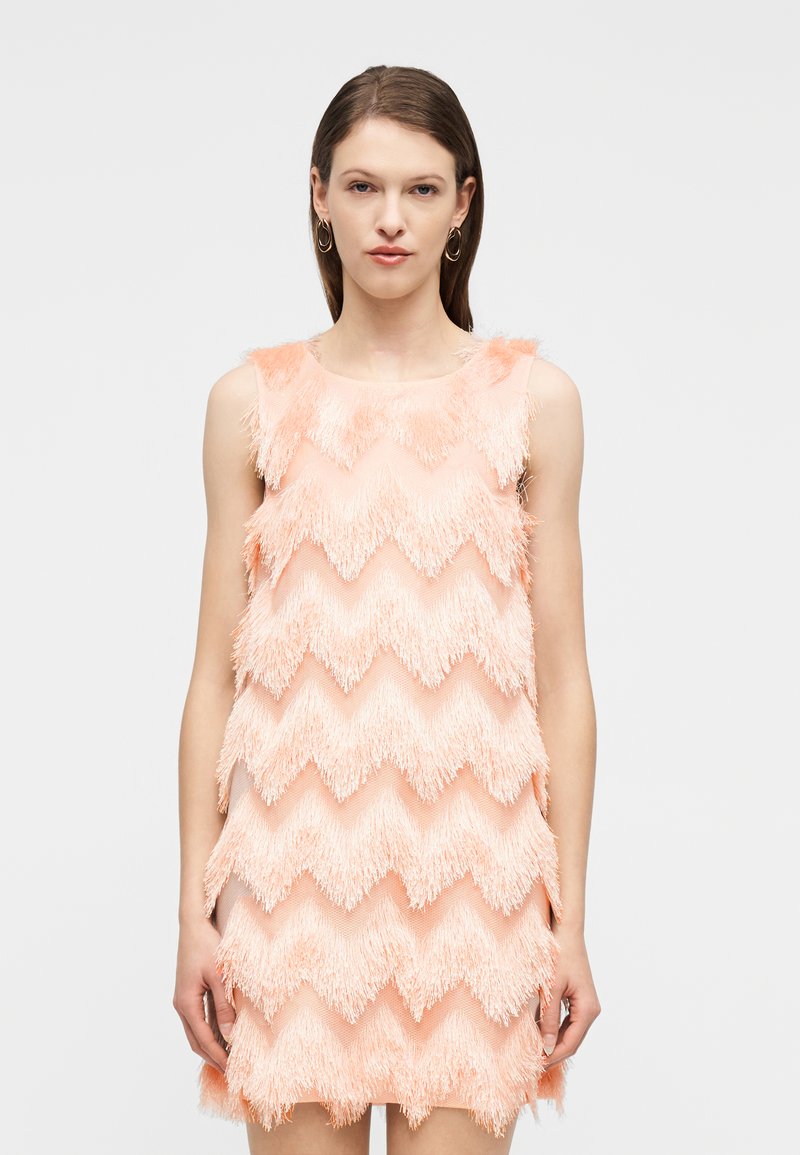 Γυναίκα που φοράει μάνικο φόρεμα σε ροδακινί απόχρωση με ανάγλυφη διακοσμητική φράντζα με ζικ-ζακ μοτίβο, στέκεται μπροστά σε απλό ανοιχτό γκρι φόντο.