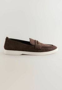 Mocassin à enfiler en daim marron avec semelle en caoutchouc blanche et détail de bride décorative sur fond neutre.