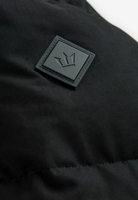 Sort polstret jakke i stof med en tekstureret, mat finish. Har et firkantet sort gummilogo-patch med en krone design.