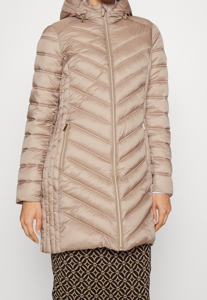 Abrigo de invierno acolchado beige con costura en forma de flecha, capucha y bolsillos con cremallera. Textura suave y cierre frontal con cremallera.