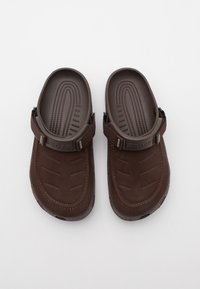 Sabots en mousse marron avec une semelle texturée, une sangle ajustable et des lignes surélevées sur la semelle intérieure pour le confort. Matériau lisse et durable.