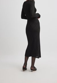 Femme de dos, vêtue d'une longue robe noire texturée, de collants noirs transparents et de chaussures à talons kitten noires dans un espace neutre.