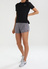 Camiseta atlética negra de manga corta con un ajuste Slim, combinada con pantalones cortos grises con aberturas laterales. Zapatillas blancas completan el conjunto.