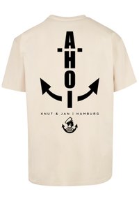 Beige T-shirt met zwarte tekst en graphics, inclusief "AHOI," pijlen en een zeemanontwerp, met daaronder "KNUT & JAN | HAMBURG."