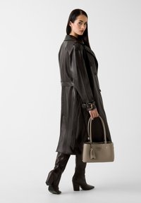 Brauner Ledermantel mit Gürtelbund, kniehohe braune Stiefel und eine graue Handtasche mit goldenen Akzenten. Glatte Textur und strukturierte Form.