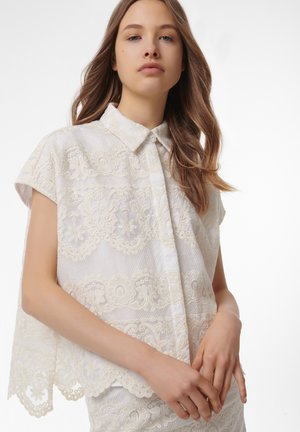Weiße Bluse aus floraler Spitze mit einem kragenförmigen Ausschnitt, kurzen Ärmeln und gewellten Kanten; aufwendige Stickdetails über den gesamten Stoff.