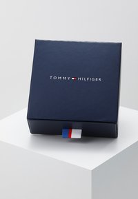 Laatikko tummansinisessä värissä, jossa on valkoinen "TOMMY HILFIGER" -logo ja punavalkoinen nauha sivussa. Sileä pinta.