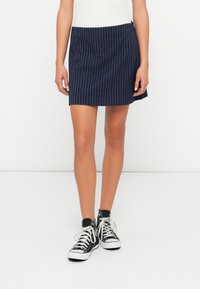 Jonge vrouw in een marineblauwe gestreepte rok, witte top en zwarte high-top sneakers, staande tegen een plain witte achtergrond.