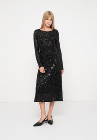 Czarna sukienka z długim rękawem z materiału wyszywanego cekinami, z dopasowanym krojem i długością do kolan. W zestawie z czarnymi butami na szpilce.