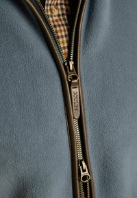 Primo piano di giacca in pile blu con cerniera rifinita in pelle marrone e camicia a quadri visibile sotto.