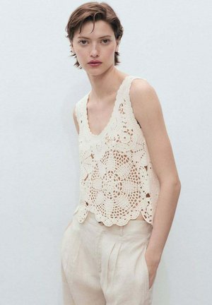 Crochet tank top in cream with floral patterns, scalloped hem, and wide straps. Paired with light beige high-waisted trousers.