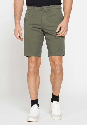 Uomo che indossa pantaloncini verde oliva, calzini neri, scarpe da ginnastica bianche e maglietta nera, in piedi davanti a uno sfondo chiaro uniforme.