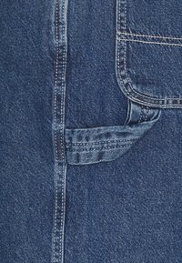 Denim fabric close-up featuring dark blue tone, contrasting stitching, and a pocket detail on the side. Textured with a smooth finish.