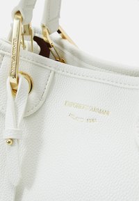 Biała torebka skórzana z żwirowym wykończeniem, ze złotymi elementami metalowymi, z wytłoczonym na złoto logo marki Emporio Armani Milano 1981.