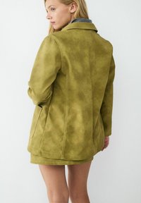 Jasnozielona, zamszowa kurtka z usztywnionym kołnierzem, bocznymi kieszeniami i widocznymi przeszyciami. Pokazana z tyłu o luźnym kroju.
