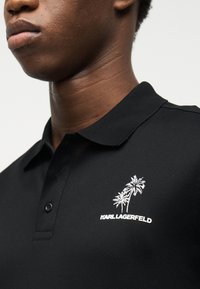 Κοντινό πλάνο προσώπου που φοράει μαύρο πόλο με δύο λευκά κεντημένα φοίνικες και την επιγραφή "KARL LAGERFELD" στο στήθος.