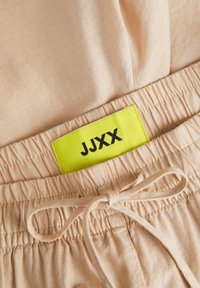 Beiga shorts med elastisk midja och snörning, med en ljusgul etikett som läser "JJXX." Mjuk tygtextur och rynkad midjedetalj.