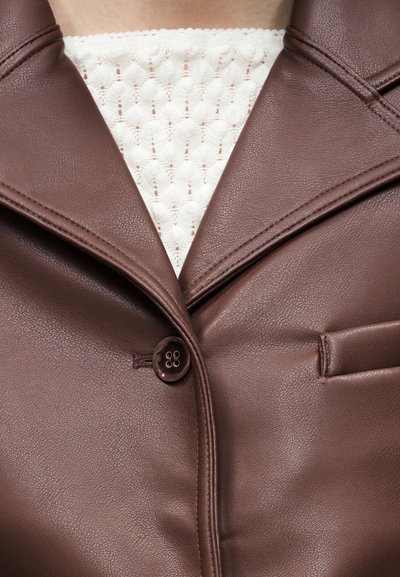 Καφέ δερμάτινο σακάκι με ένα μόνο κουμπί, φορεμένο πάνω από λευκό πλεκτό τοπ με υφή, εμφανίζεται από το στήθος και πάνω.