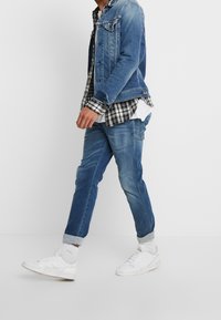 Denimjacka i mellanblått, bärs över en svartvit rutmönstrad flanellskjorta. Ljusa jeans med uppvikta mudd och vita sneakers.