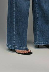 Jeans en denim bleu à jambe large avec un ourlet effiloché, associés à des sandales noires à bout ouvert présentant un design épuré et un petit talon carré.