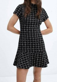 Mujer con un vestido de cuadros en blanco y negro de manga corta y dobladillo acampanado, de pie con las manos detrás de la espalda sobre un fondo blanco.