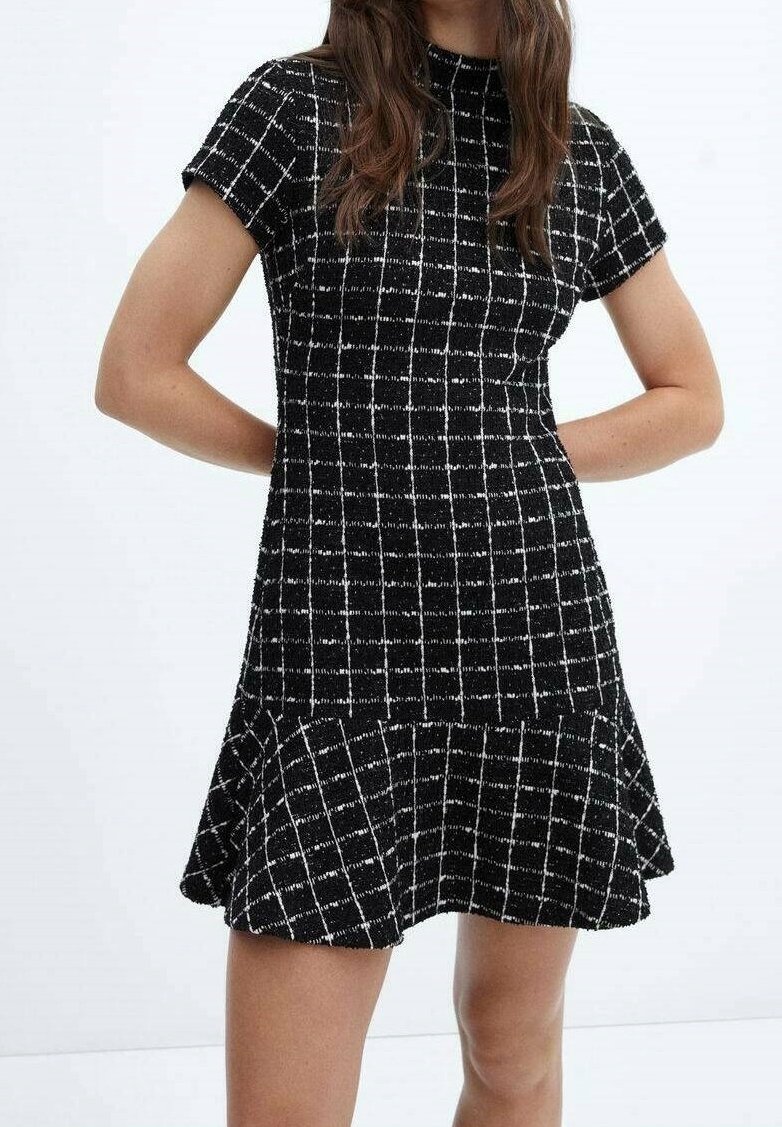 Mujer con un vestido de cuadros en blanco y negro de manga corta y dobladillo acampanado, de pie con las manos detrás de la espalda sobre un fondo blanco.