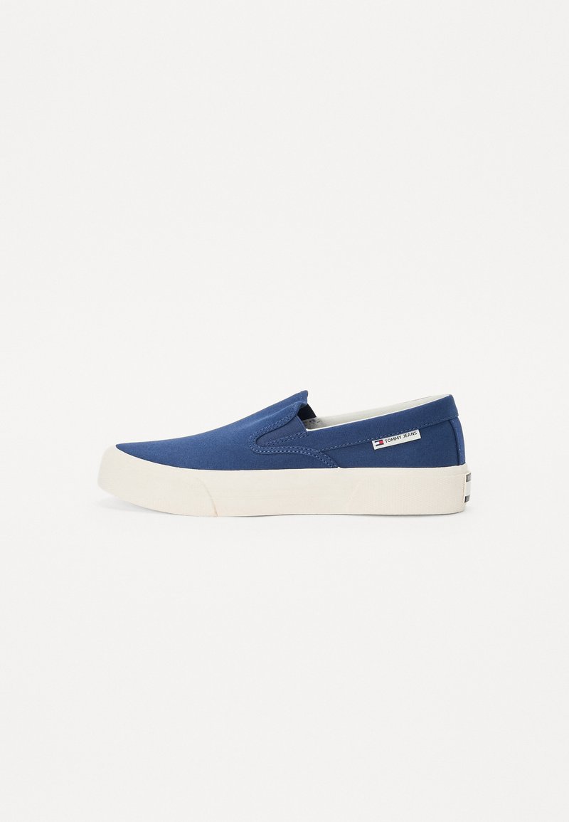 Zapatillas de lona azul sin cordones con una suela de goma blanca. Presentan paneles elásticos a los lados y una pequeña etiqueta con el logo. Textura suave, diseño de punta redondeada.