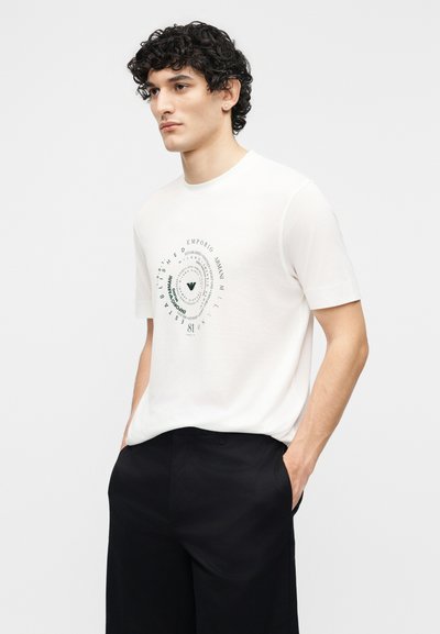 Νέος άνδρας με σγουρά μαλλιά, που φοράει λευκό κοντομάνικο μπλουζάκι με κυκλικό μαύρο κείμενο και μαύρο παντελόνι, με τα χέρια στις τσέπες, στραμμένος προς τα αριστερά.