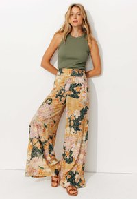 Pantalons larges à fleurs en jaune, rose et vert avec une taille smockée. Associés à un haut en olive et des sandales marron.