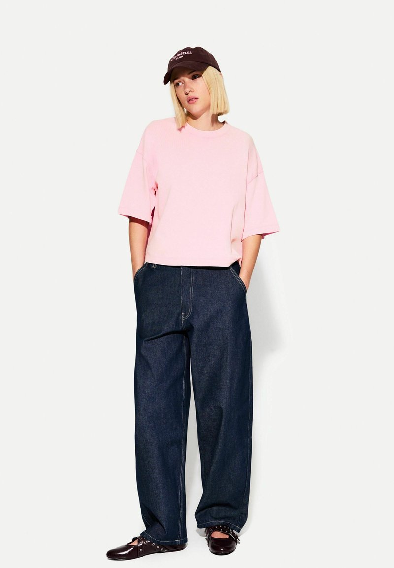 Růžové zkrácené triko, volné tmavé denimové kalhoty, hněvý kšilt s bílým textem a tmavé boty s dekorativními prvky. Neformální outfit.