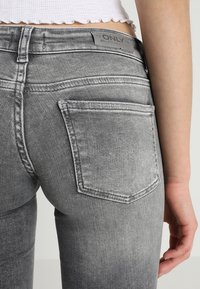 Jeans de mezclilla gris con una textura desgastada, diseño de cinco bolsillos y un bolsillo trasero visible. Presenta una etiqueta de marca cerca de la cintura.
