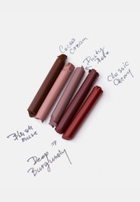 Cinq bâtons de rouge à lèvres cassés dans des tons marron et rouge, étiquetés Cocoa Cream, Blush Muse, Dusty Rose, Deep Burgundy et Classic Cherry, posés sur une surface blanche.
