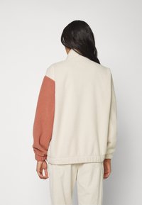 Chaqueta de forro polar con un cuerpo blanco y una manga de color óxido, cuello alto, puños elásticos y un corte relajado con una superficie texturizada.