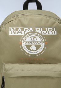Zaino verde oliva con superficie testurizzata, dotato di un logo bianco, grafica circolare e una tasca frontale con zip. Spallacci neri in alto.