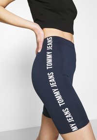 Marina blå cykelshorts i elastiskt material med vit text "TOMMY JEANS" vertikalt längs sidan, med en slät och åtsittande design.