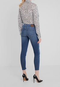 Los pantalones vaqueros ajustados azules presentan una cintura de altura media y bolsillos traseros, combinados con una blusa de manga larga transparente y estampada, y tacones altos negros.