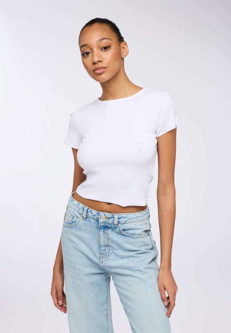 T-shirt bianco croppato con maniche corte, realizzato in tessuto a costine. Presenta dettagli decorativi sulla spalla. Abbinato a jeans azzurri chiari.