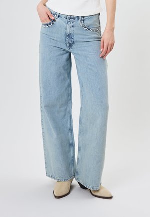 Person trägt hellblaue Jeans mit weitem Bein und gezackter weißer Naht an den Fronttaschen, weißes Oberteil und beige spitze Schuhe.