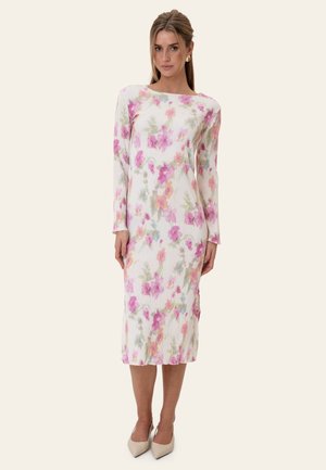 Vrouw draagt een witte midi-jurk met lange mouwen en een roze en paarse bloemenprint, gecombineerd met beige puntige hakken.