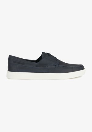 Chaussures en toile bleu marine à enfiler avec une finition texturée, semelle en caoutchouc blanc et couture décorative le long des bords. Présente un laçage minimal.